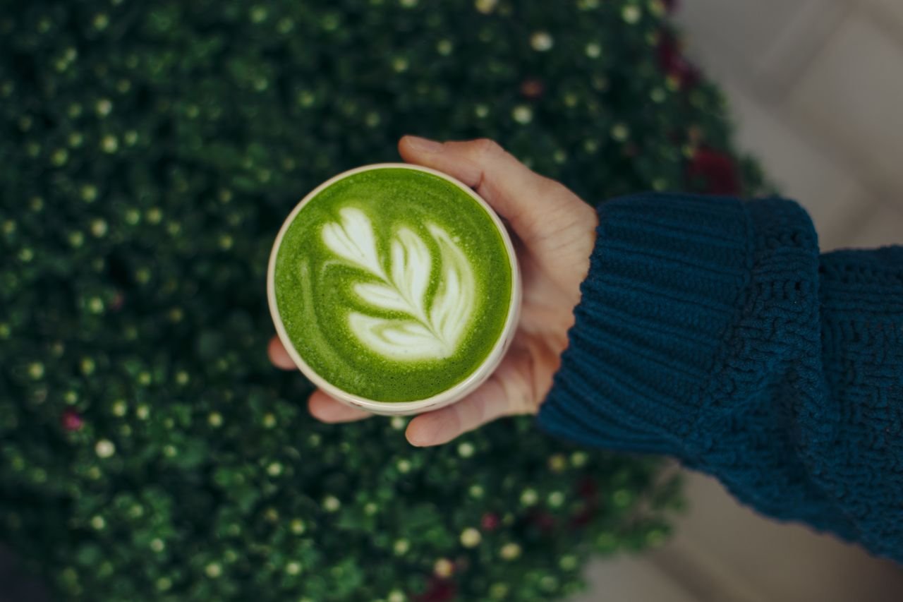 Home A close-up of a matcha latte with latte art, held by a hand in a cozy knitted sweater, against a blurred green background.