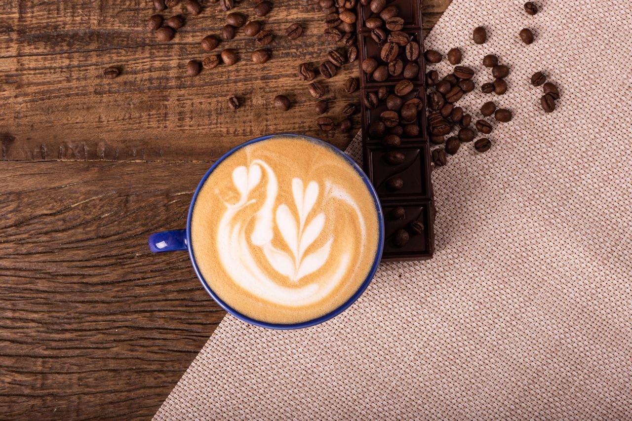 Home Overhead view of a latte with swan latte art, surrounded by coffee beans and chocolate on rustic wood.